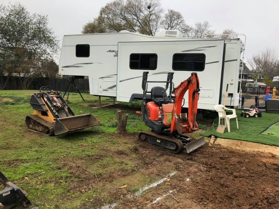 Two compact excavators beside a white RV on a muddy campsite, one digging soil
