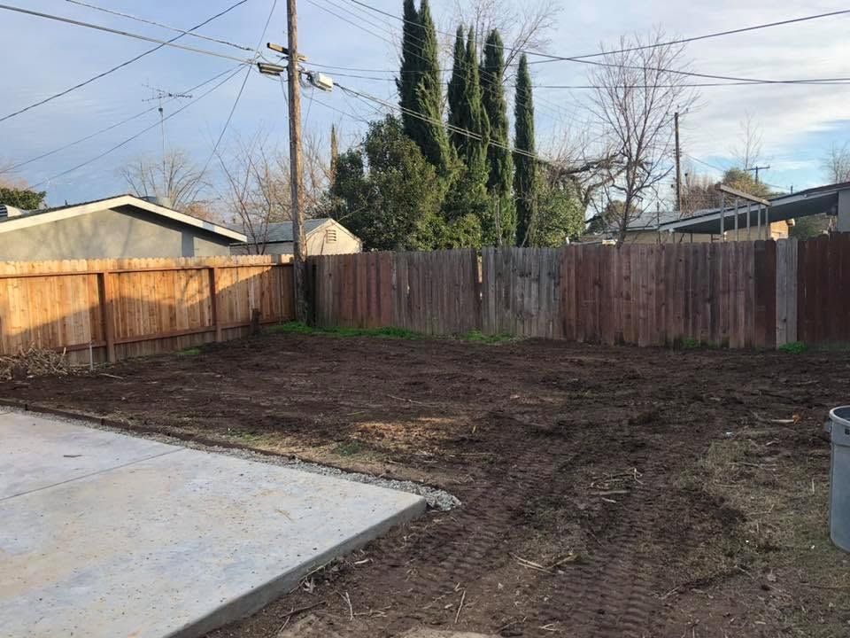 Backyard with bare dirt, wooden fence, concrete slab, and tall trees under a cloudy sky