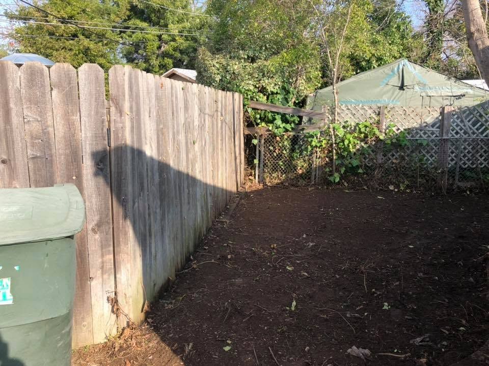 Wooden fence beside a bare yard with a green trash bin and a gazebo in the background