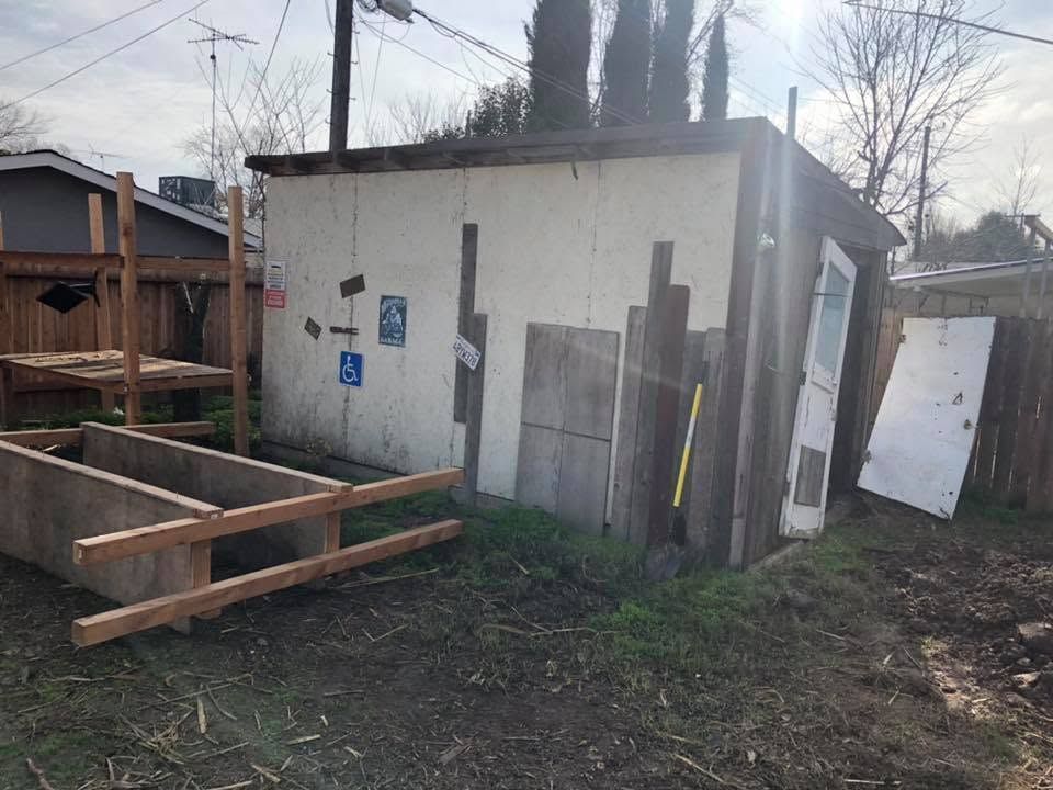 Small weathered shed with boarded side, wooden planter frame, and a white door in a muddy yard