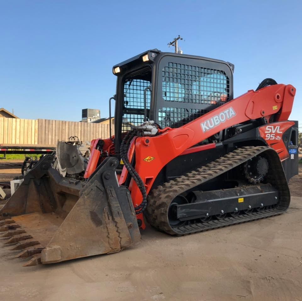 Red Kubota tracked skid steer with front bucket, parked on a dirt lot under a blue sky.