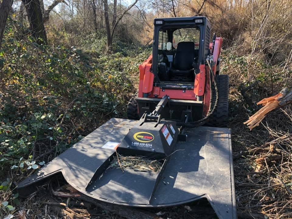 Red skid steer pushes brush with a grapple on a wooded path, clearing debris.