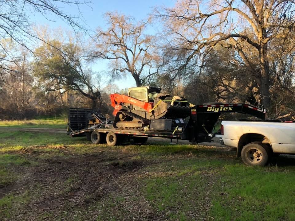 White truck towing a trailer with an orange skid steer across a grassy field at sunset