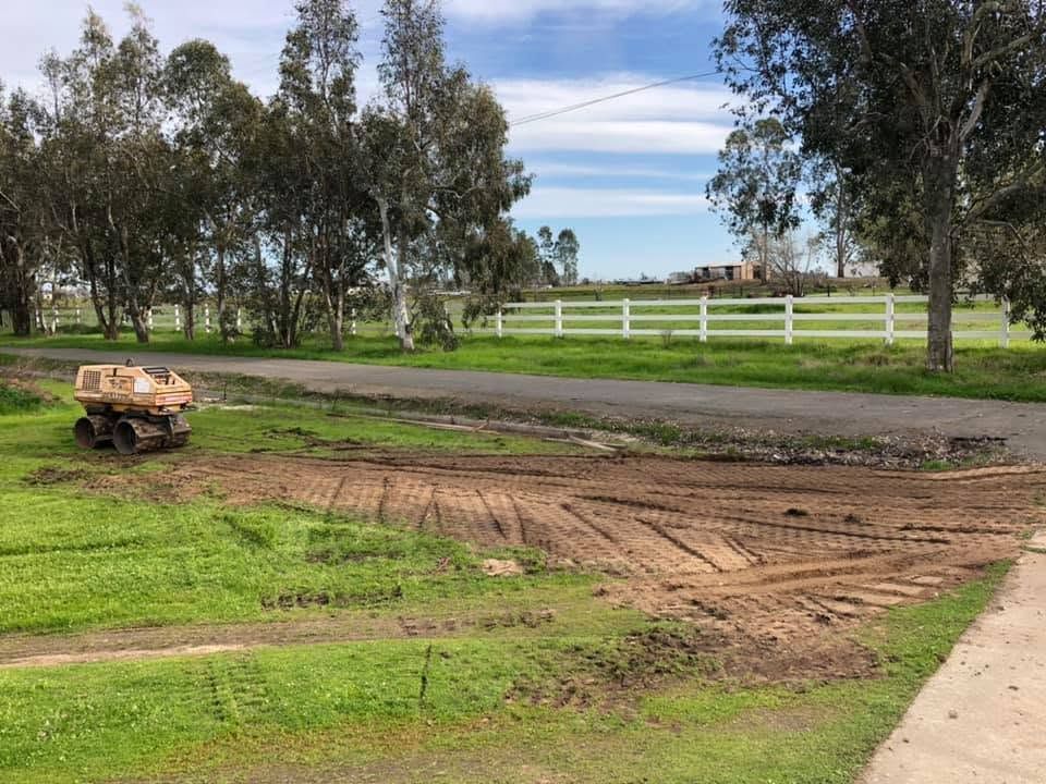 A dirt road under construction with a small dump cart, grassy field, white fence, and trees in the background.