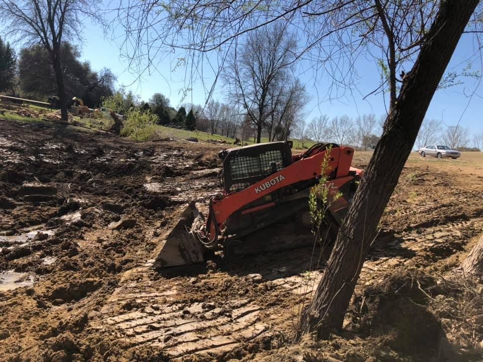 Red skid steer clearing dirt on a muddy hillside near bare trees under a blue sky.