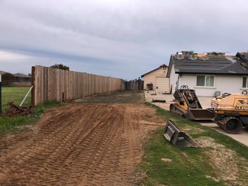 Dirt driveway beside a fenced house with a construction trailer and excavator under cloudy sky