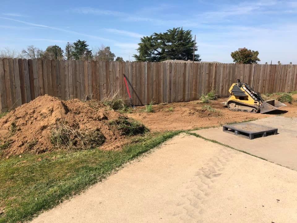 Backyard with dirt pile, yellow mini excavator, wooden fence, and concrete patio edge