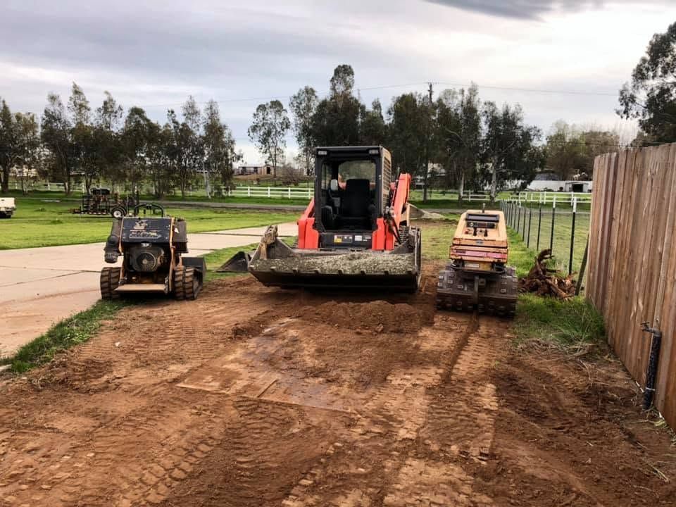 Two skid steers and a front loader on a dirt lot beside a fence and trees