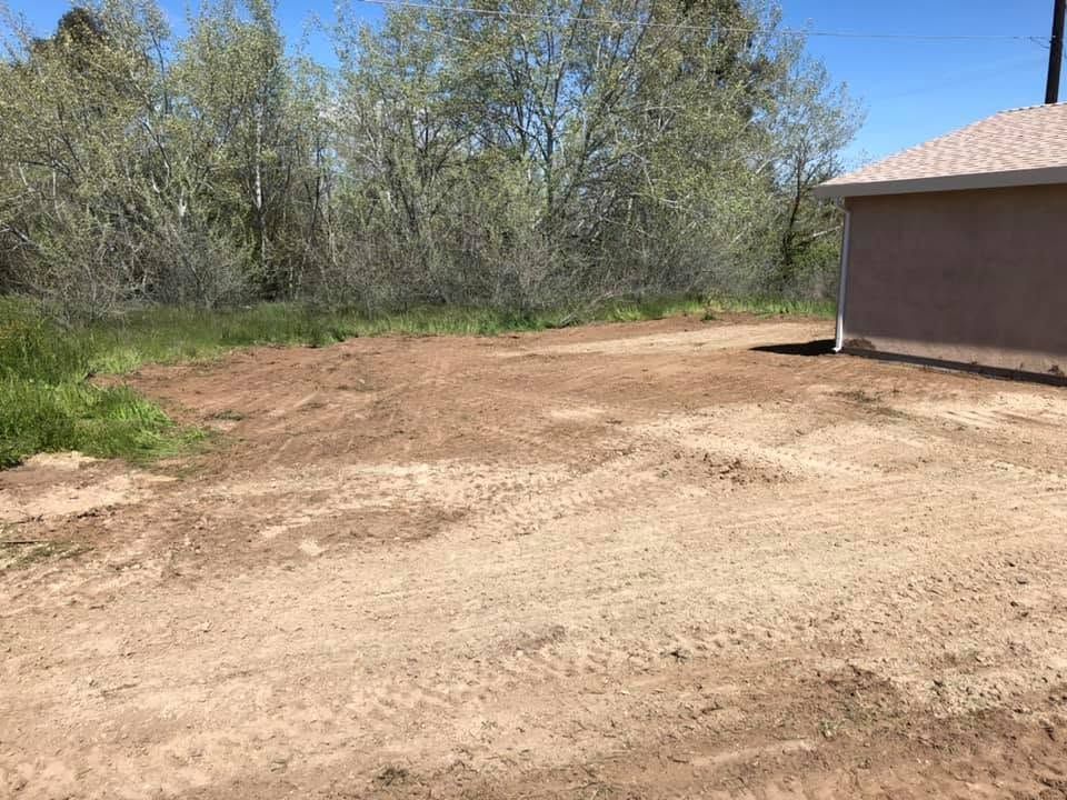 Dirt clearing beside a tan house, bordered by trees under a bright blue sky.