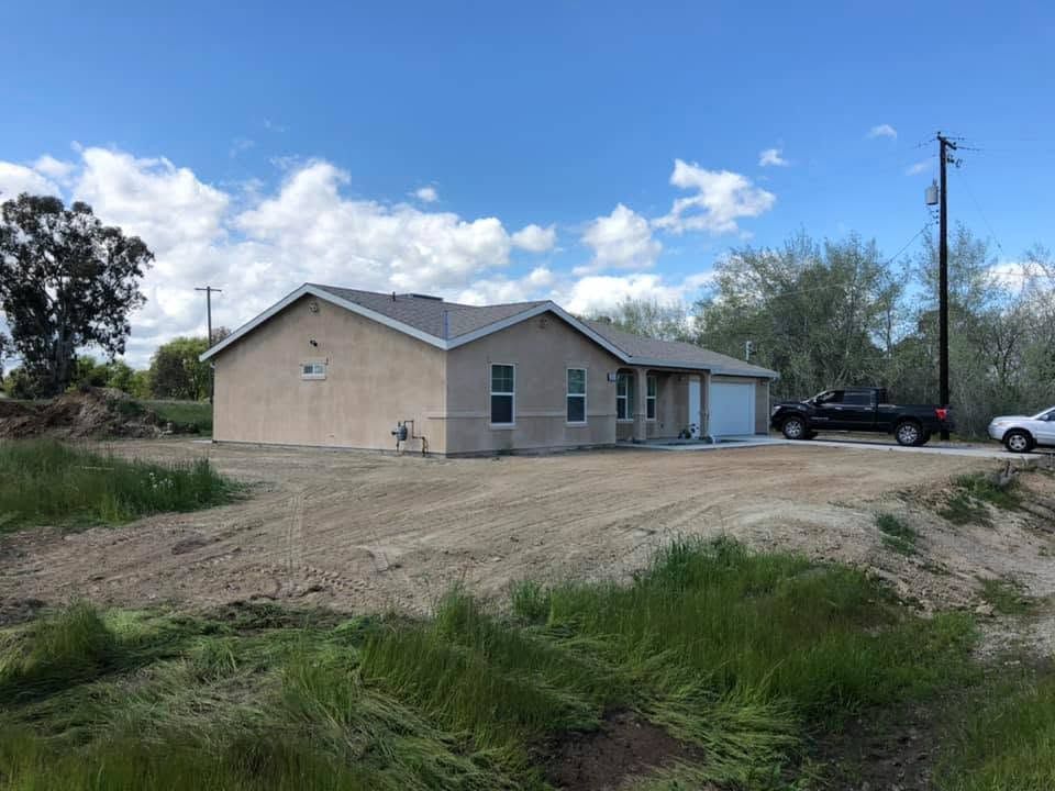 Single-story beige house on a dirt lot under a blue sky, with parked vehicles nearby.