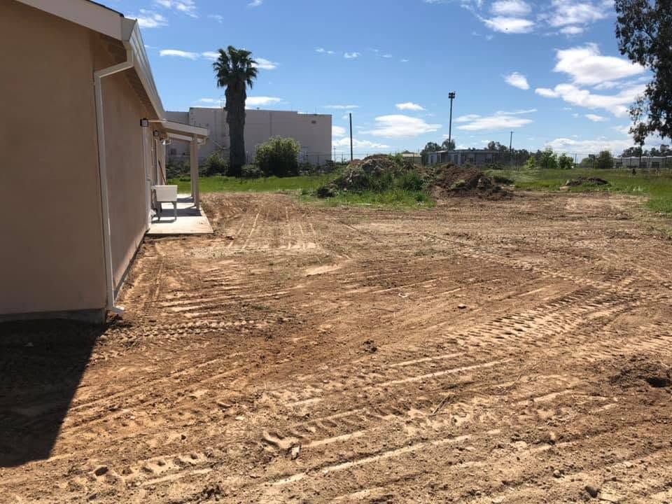 Dirt lot beside a tan building under a blue sky, with palm trees and construction debris in the distance