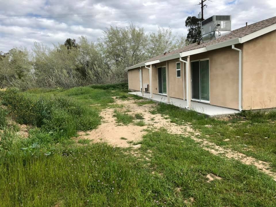 Single-story beige house with green doors beside a grassy dirt path under a cloudy sky
