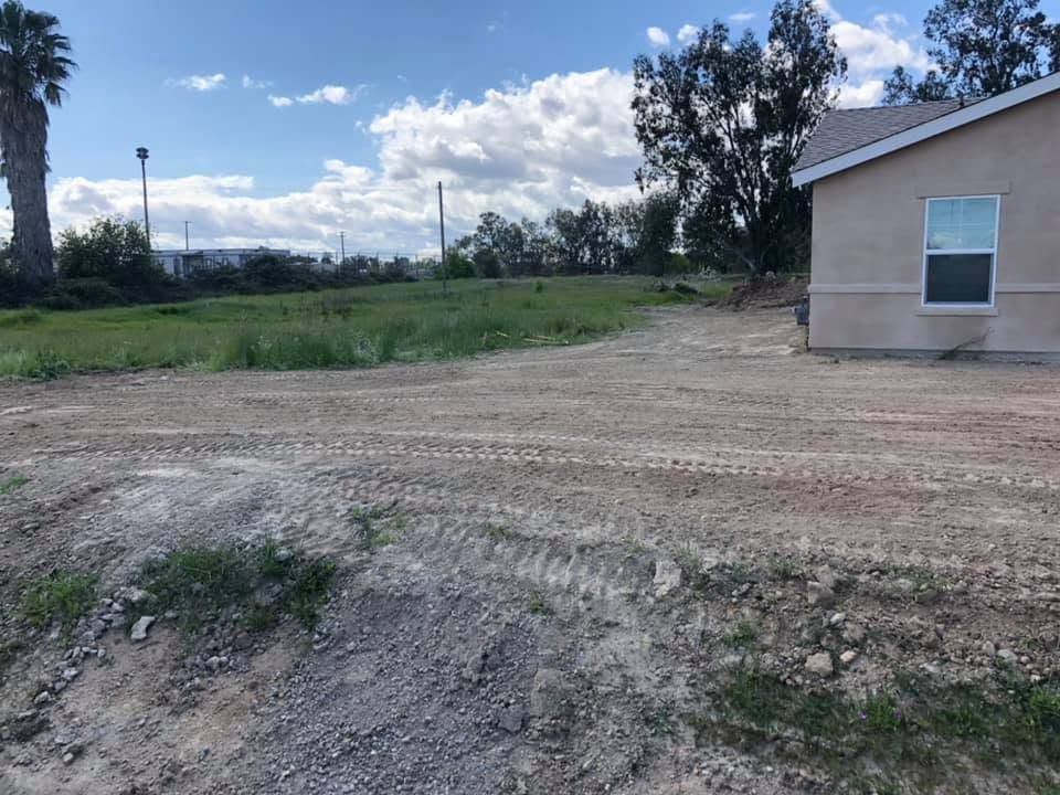 Gravel driveway beside a beige house, opening to a grassy field under a cloudy blue sky.