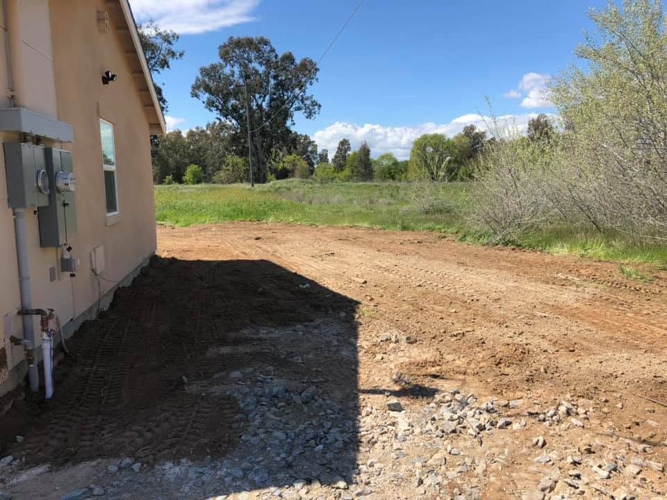 House exterior beside a dirt driveway, opening to a grassy field under a clear blue sky
