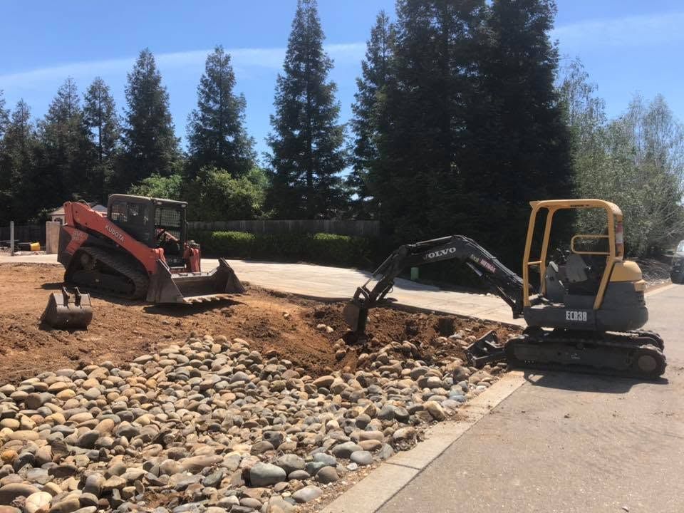 Two excavators working on a rocky dirt path beside a paved road with tall trees in the background