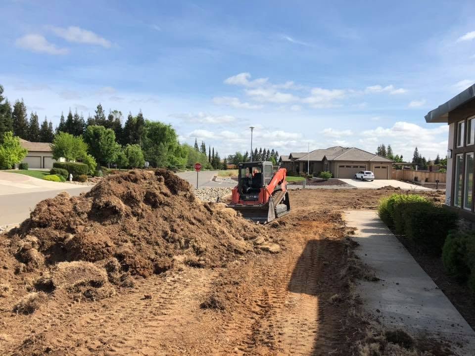 Construction site with excavator moving dirt beside a residential sidewalk under a blue sky
