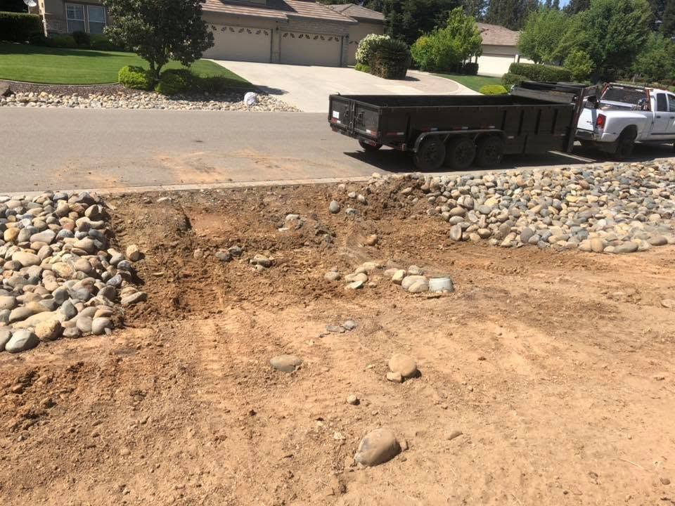 Construction dump trailer parked beside a rocky retaining wall on a dirt lot by a suburban street.
