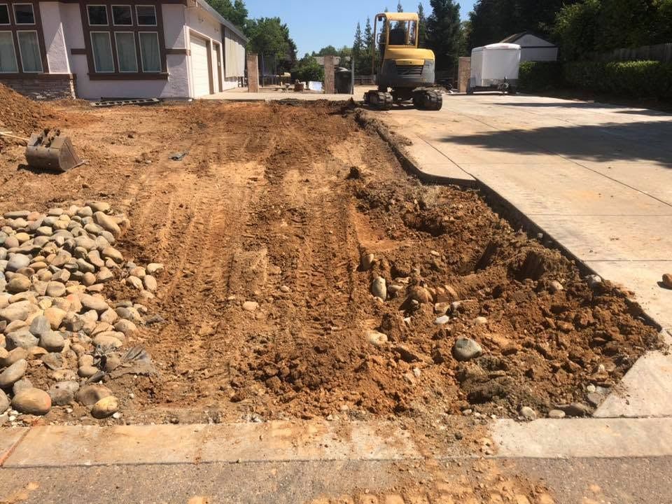 Road construction excavator grading dirt beside a residential driveway and sidewalk