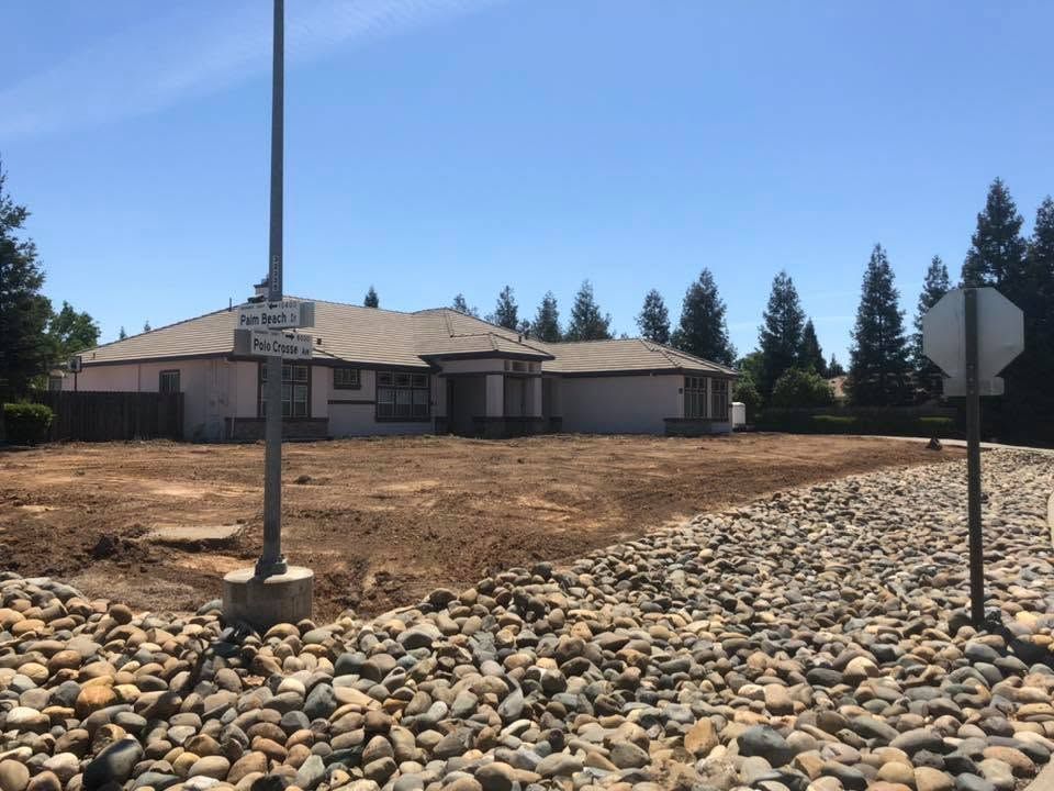 Single-story house on a dry lot with rocks, lamp post, and trees under a clear blue sky