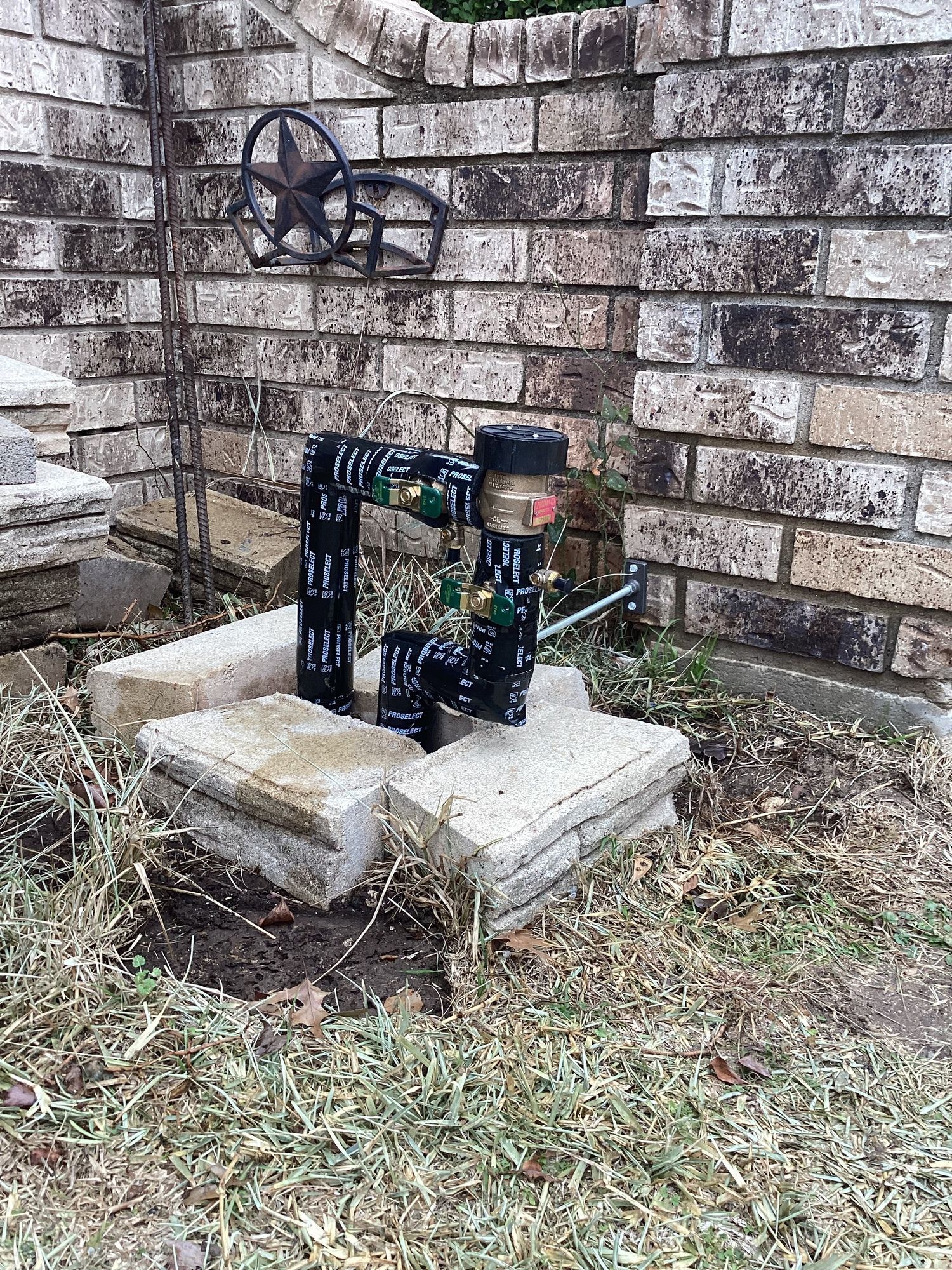 A water meter is sitting in the grass in front of a brick wall.