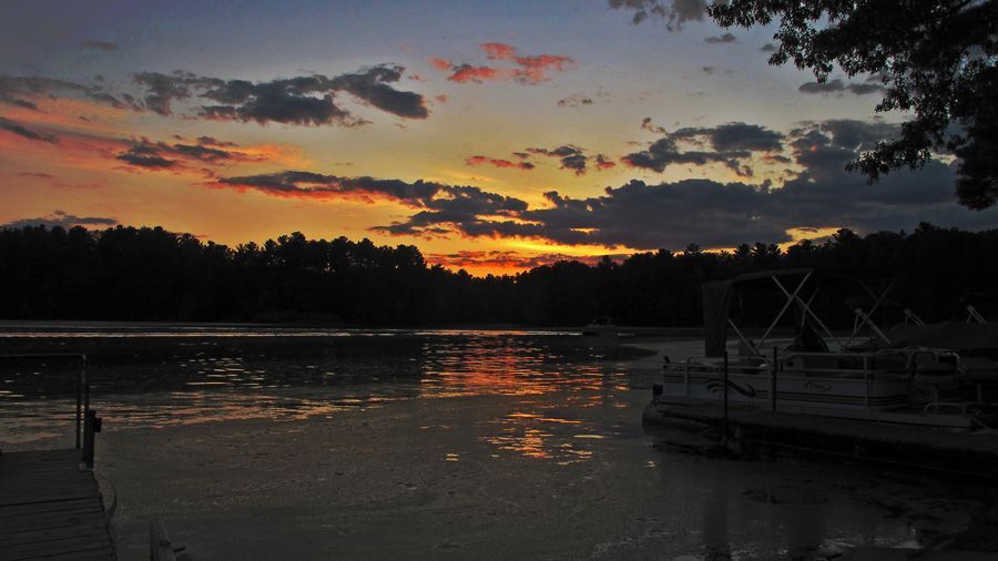 Sunset over a calm lake with orange and pink clouds, silhouetted trees, and docked boats at right