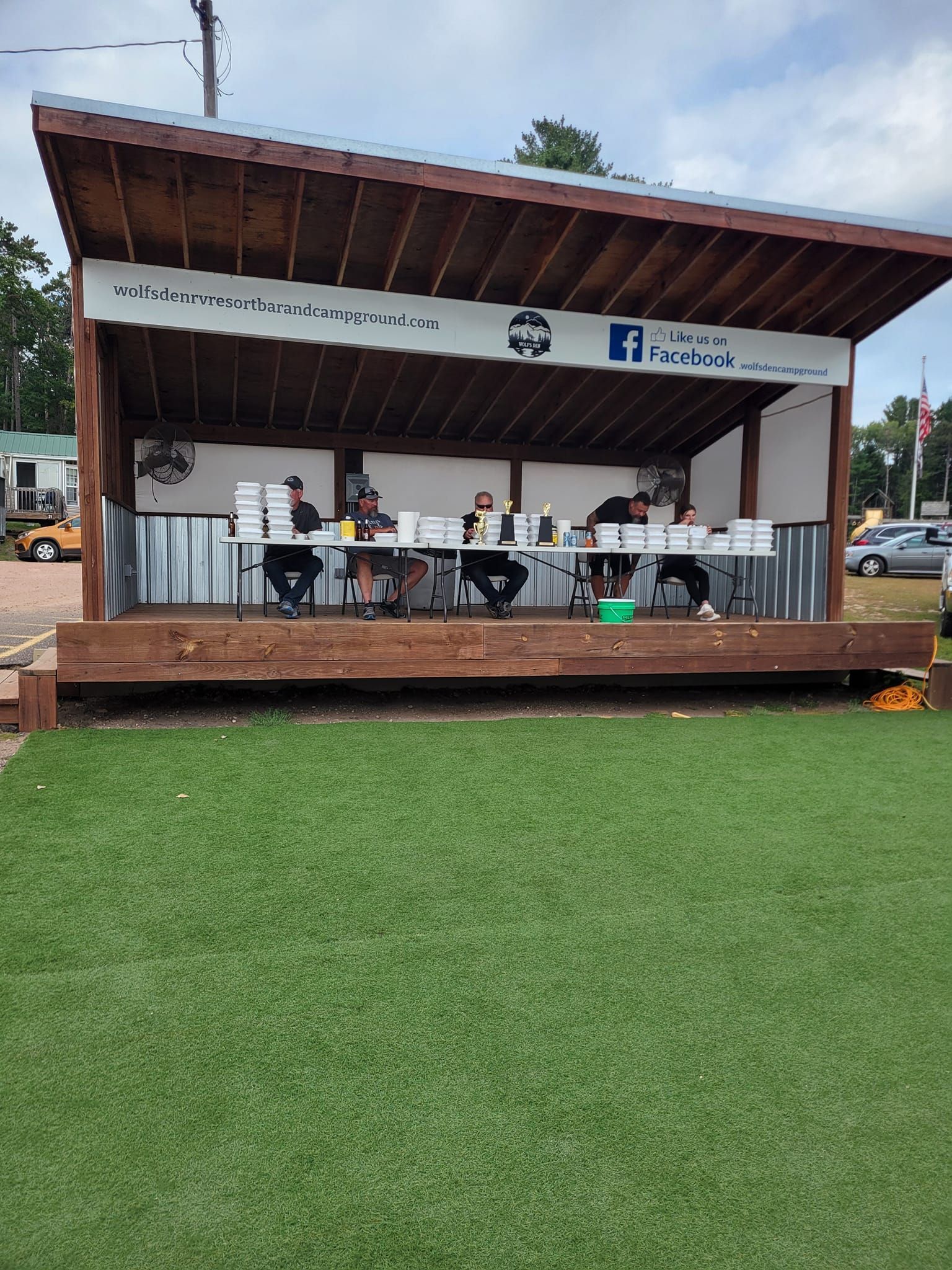 Open-air café kiosk on a wooden deck beside a grassy field, with tables and chairs under a sloped roof