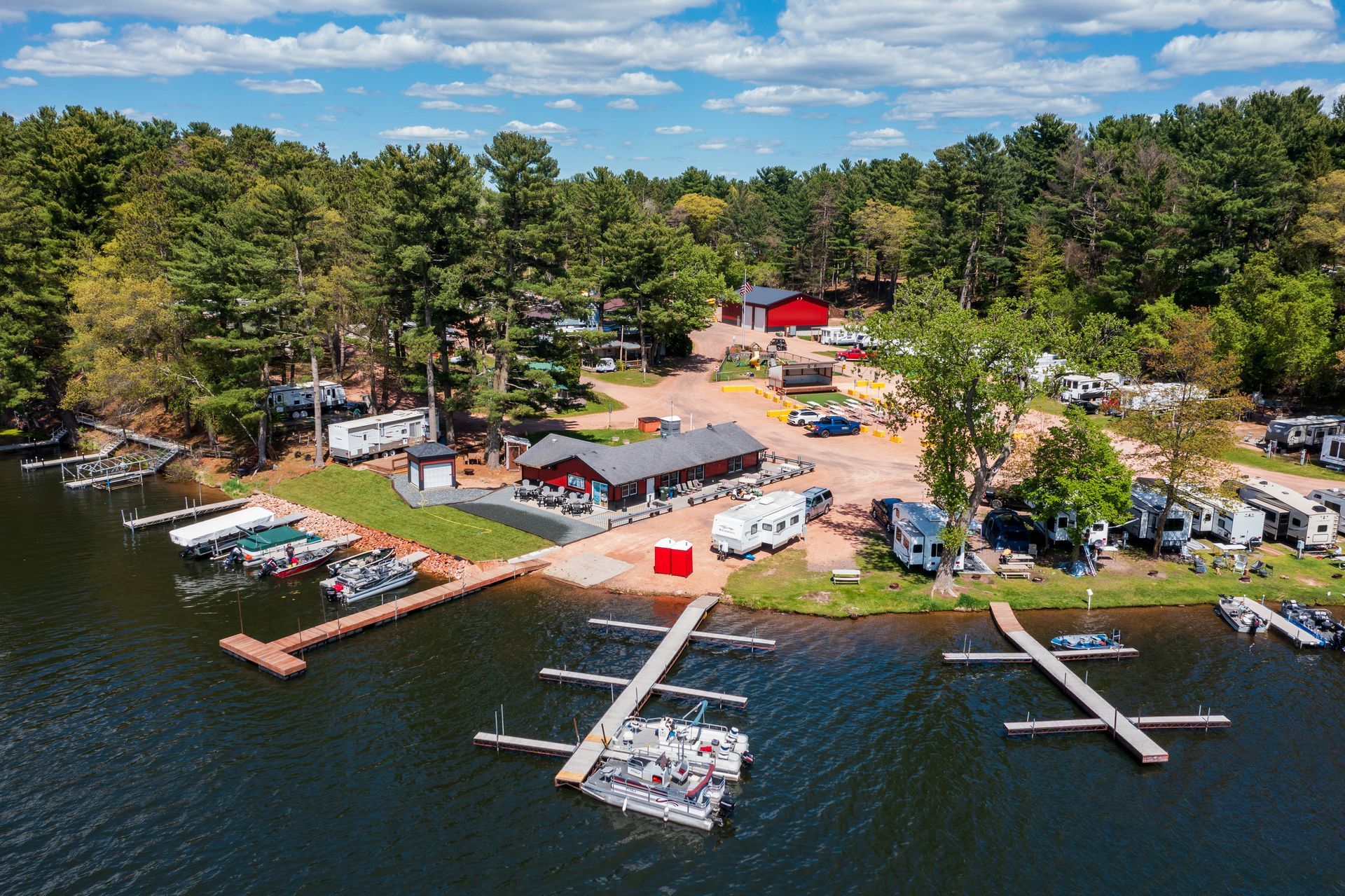 Aerial view of a lakeside marina with docks, boats, cabins, and trees under a blue sky