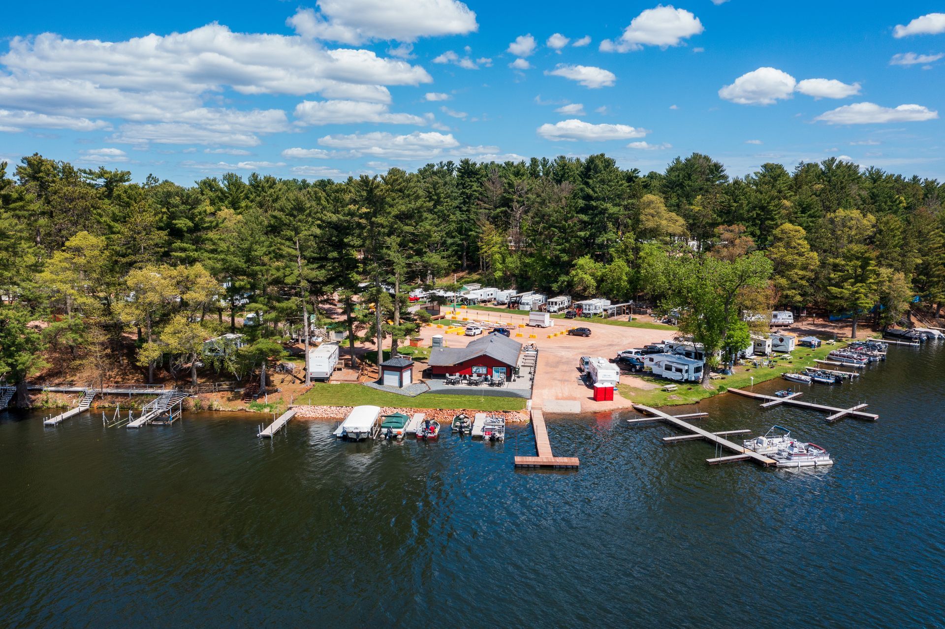 Aerial view of a lakeside campsite with docks, boats, tents, and trees under a blue sky