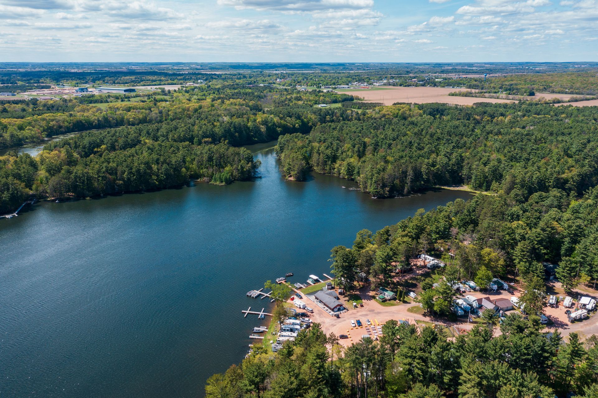Aerial view of a forested lake with a sandy beach and parked cars under a blue sky.