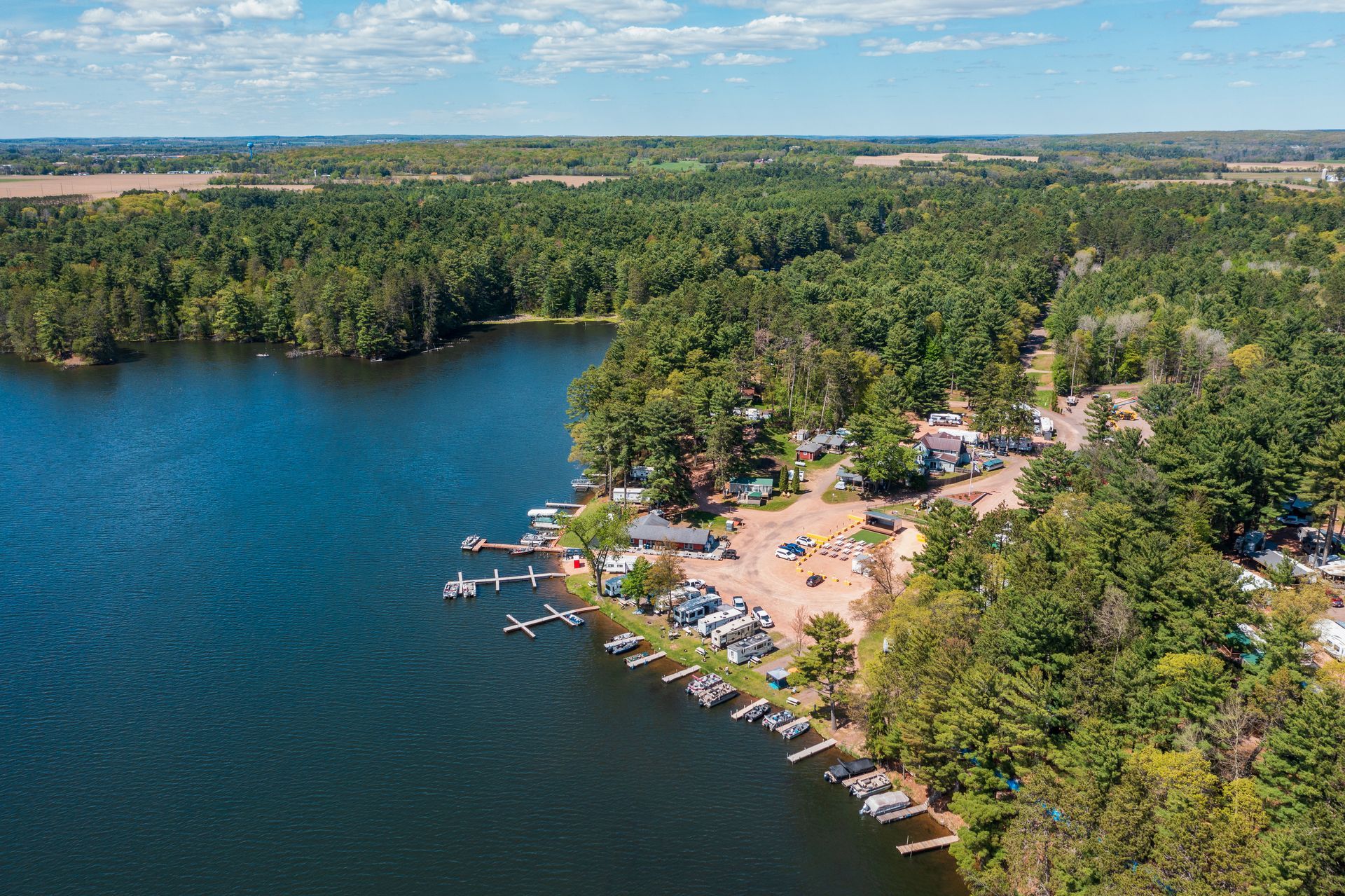 Aerial view of a lakeside beach with docks, boats, and a wooded shoreline under a blue sky.