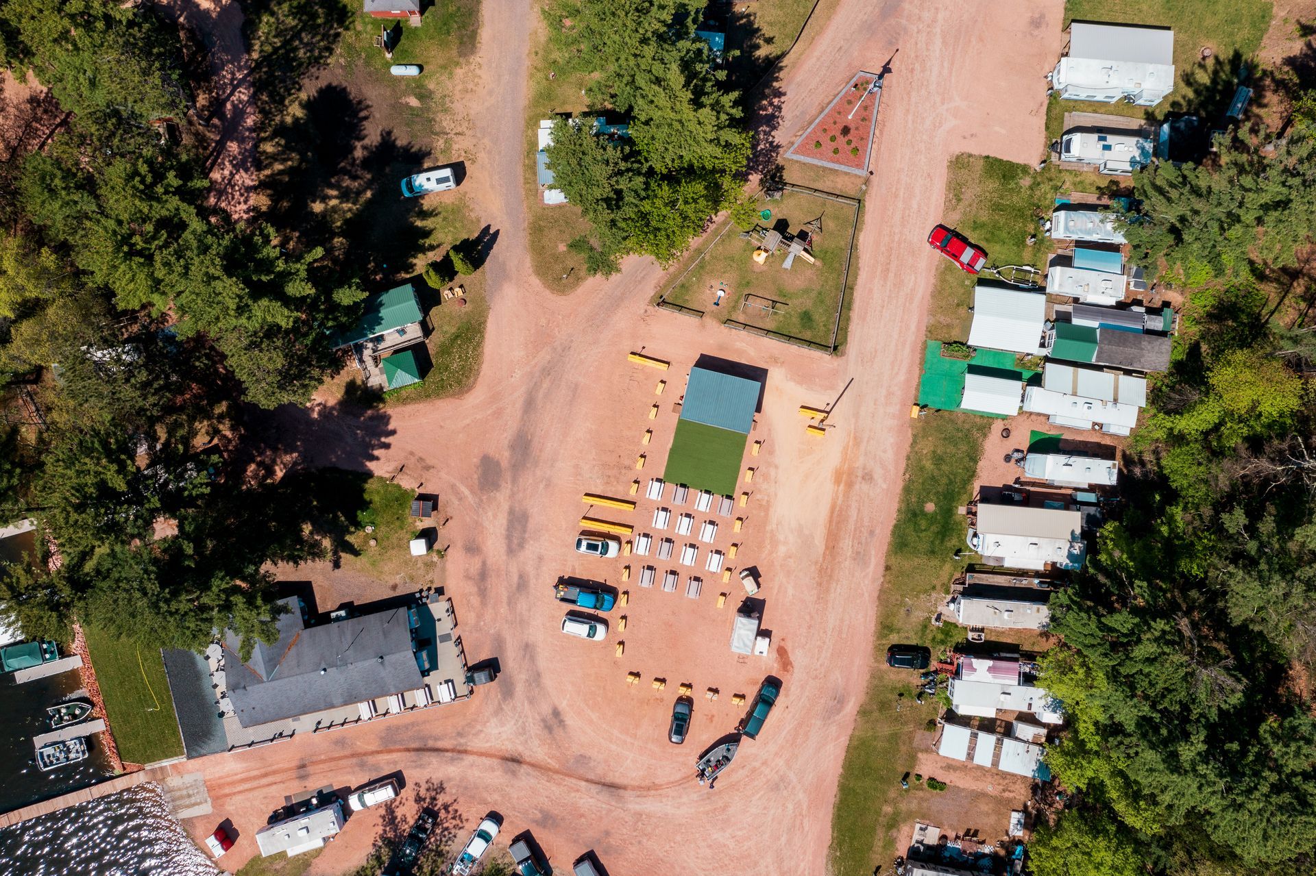 Aerial view of a dirt campsite with tents, parked vehicles, and people gathered among trees.