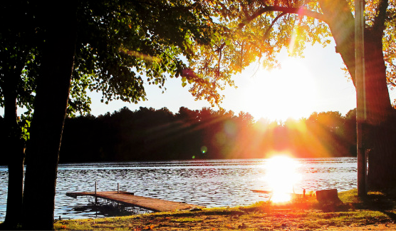 Sunset over a lake with trees silhouetted in the foreground and sunlight reflecting on the water.