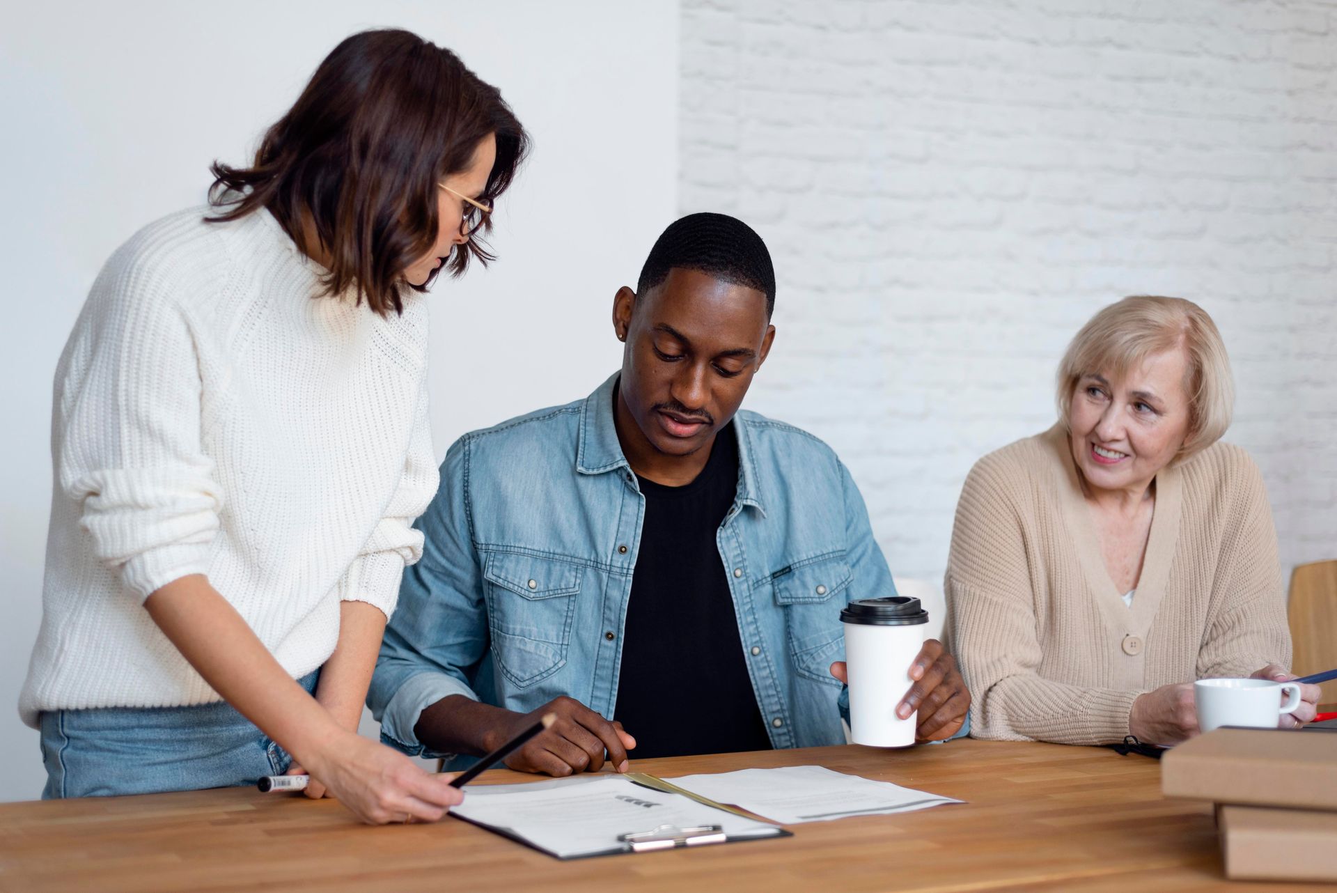 Three people review documents at a table, with one pointing and another holding a coffee cup.