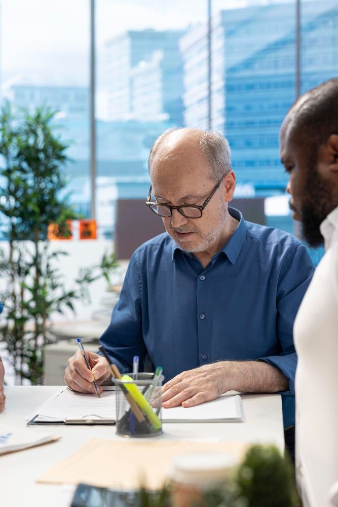 People collaborating at a bright office table, one man writing on paper with city buildings behind them