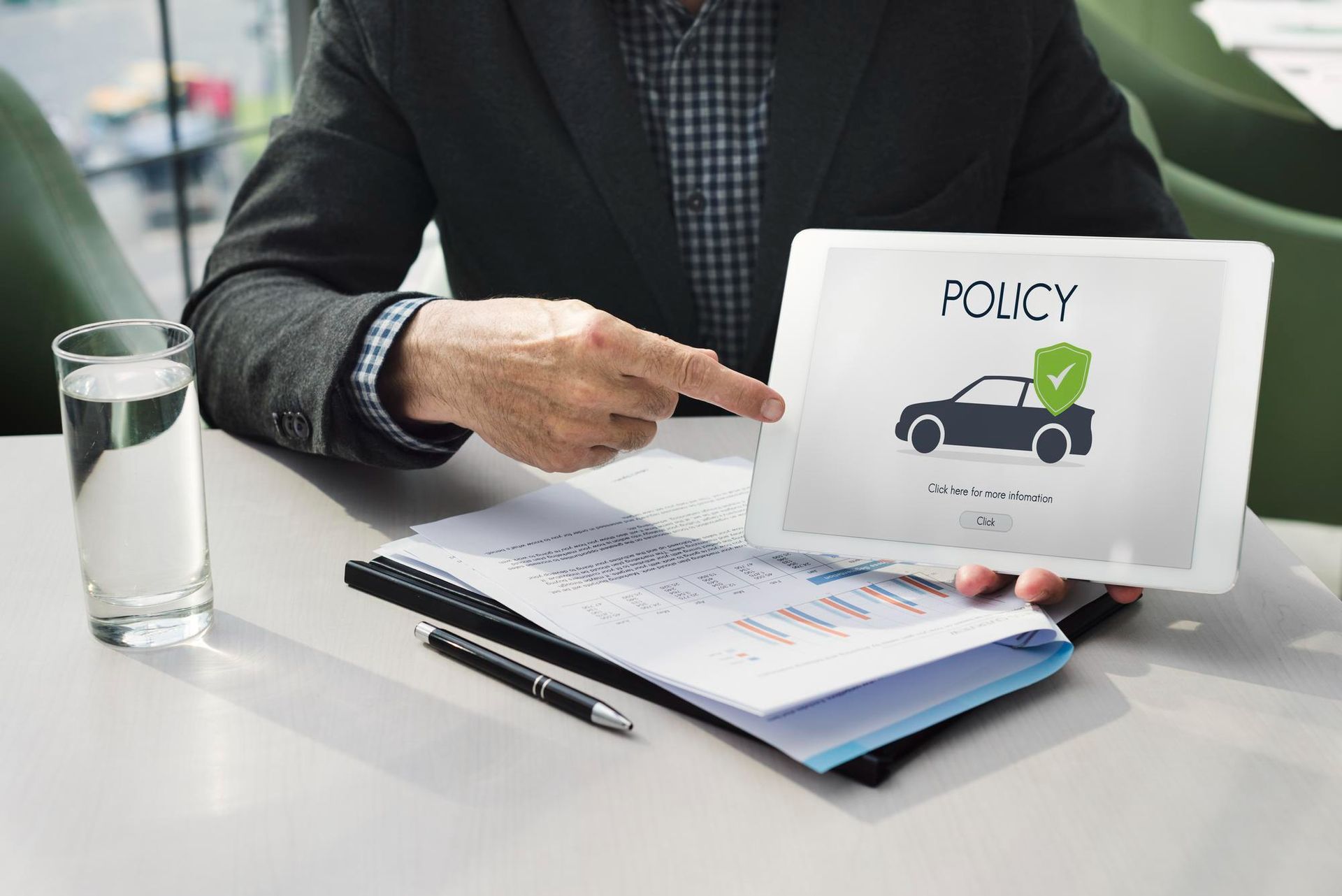 Person pointing at a tablet labeled “POLICY” beside a notebook and glass of water on a desk