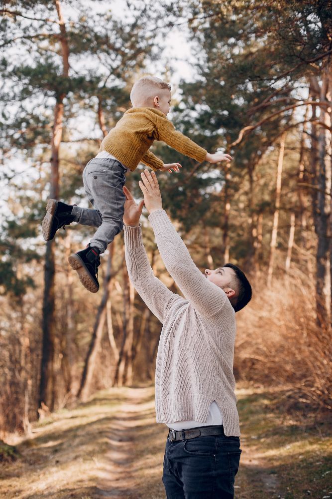 Adult lifting a child into the air on a sunlit forest path