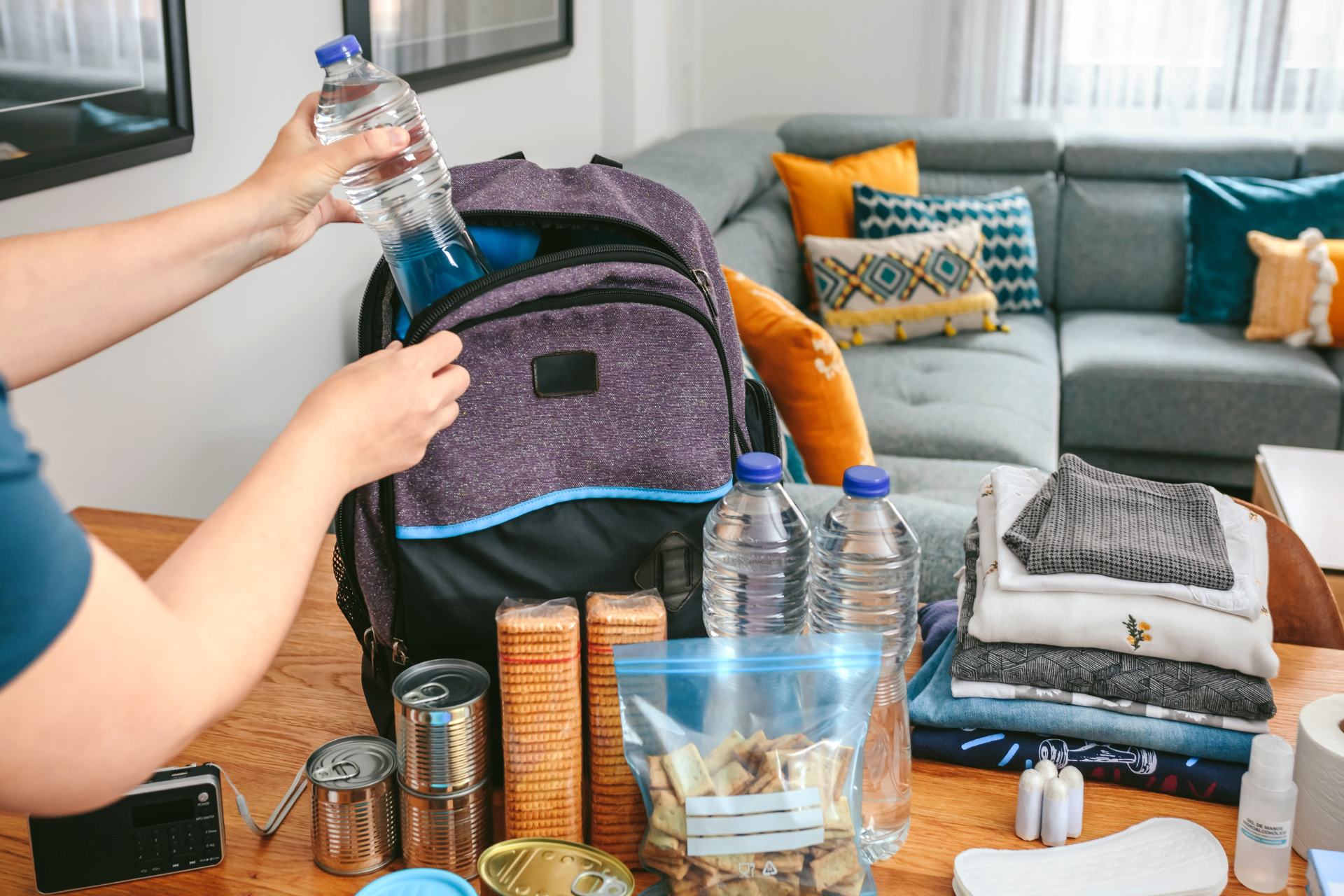 Person pouring water into a purple backpack in a living room with a couch and table of snacks