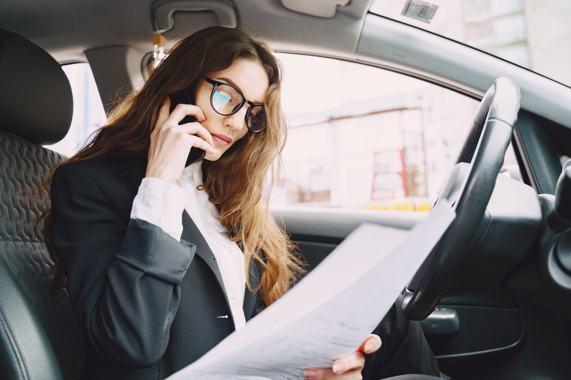 Woman in glasses reading papers and talking on a phone inside a car