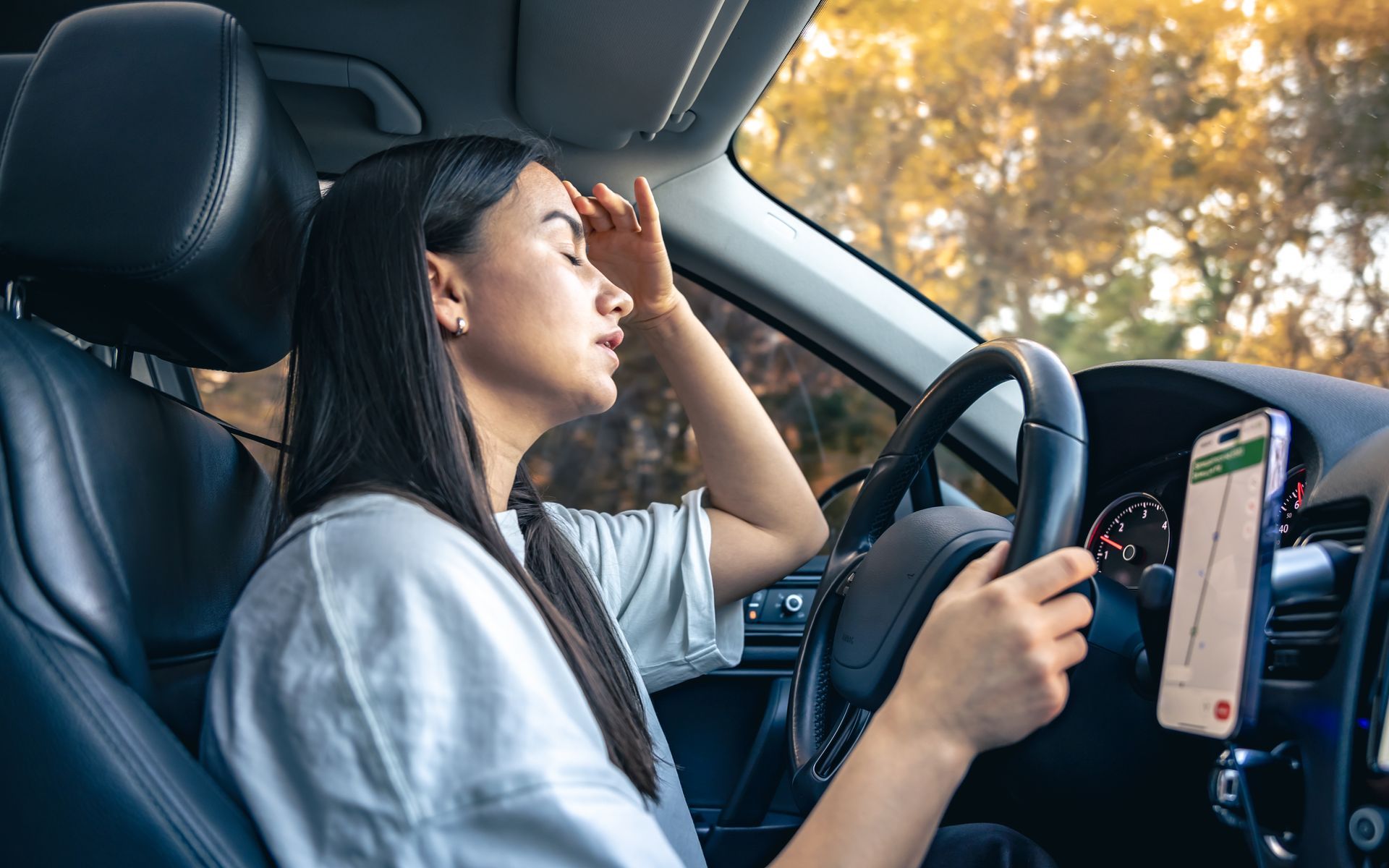 Driver in a car with one hand on the wheel and the other shielding their eyes from sunlight, using a phone mount on dashboard