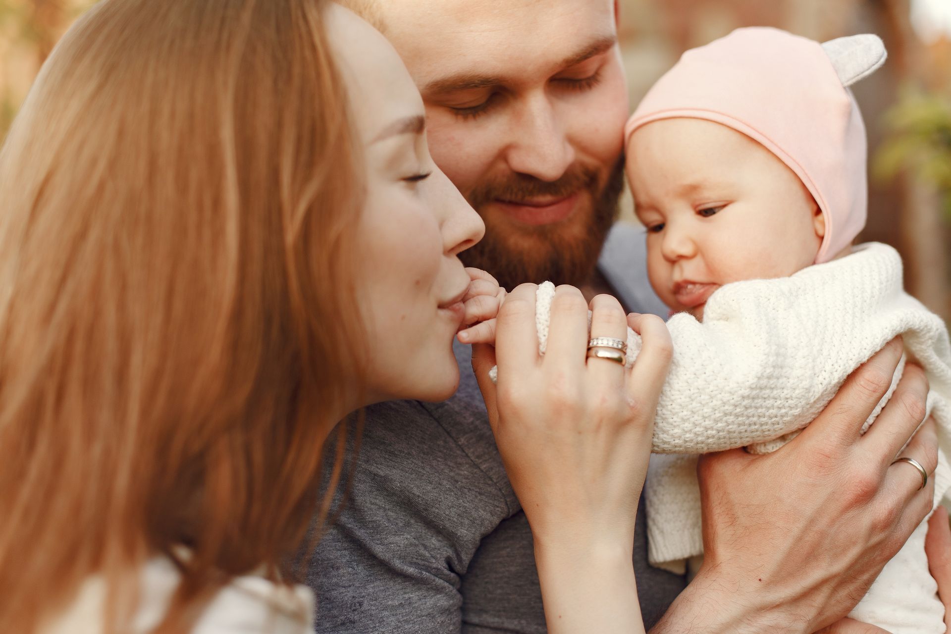 A couple cuddling a baby in a pink hat, with the mother kissing the baby’s hand.