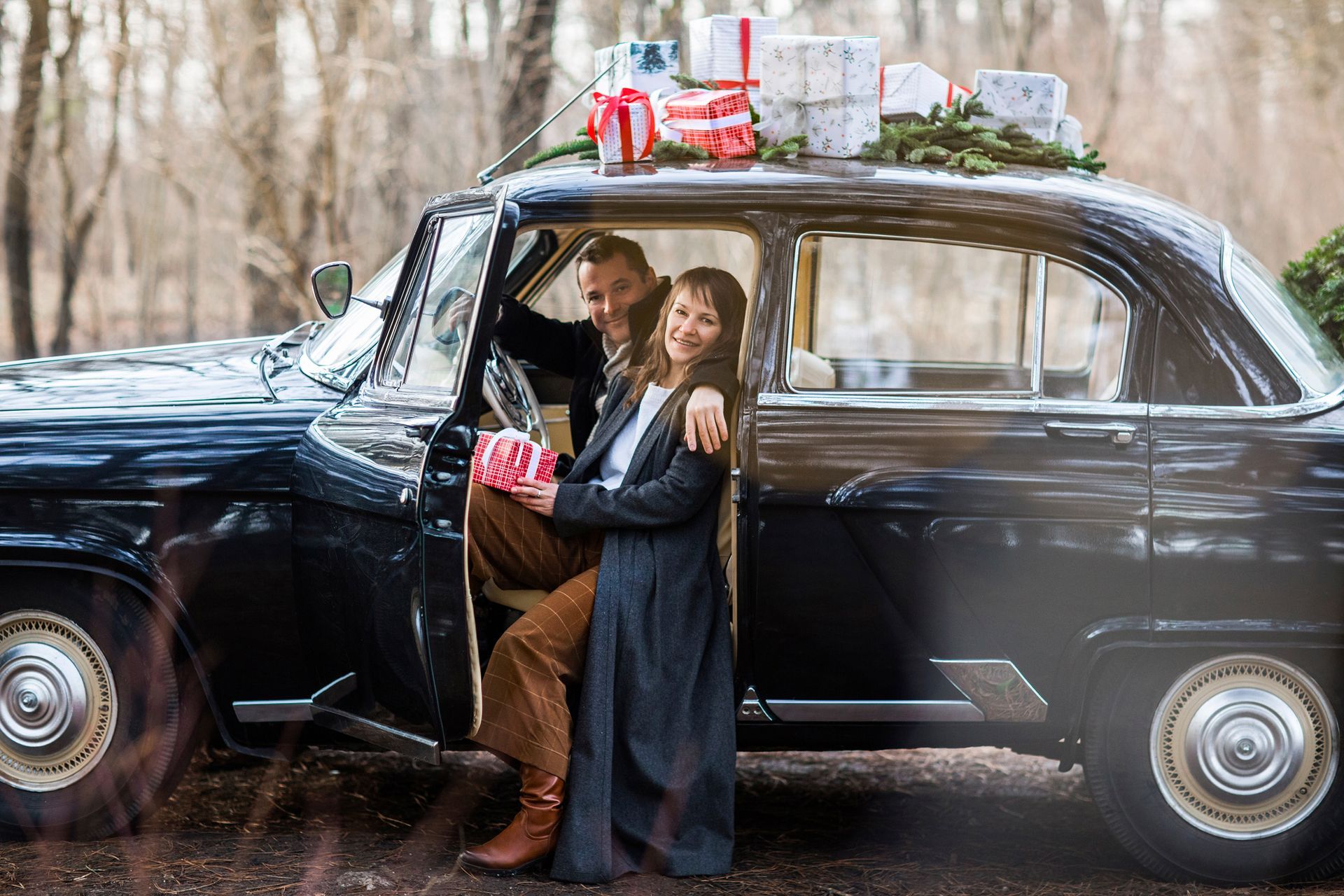 Two children by a black vintage car decorated with gifts in a winter woodland setting