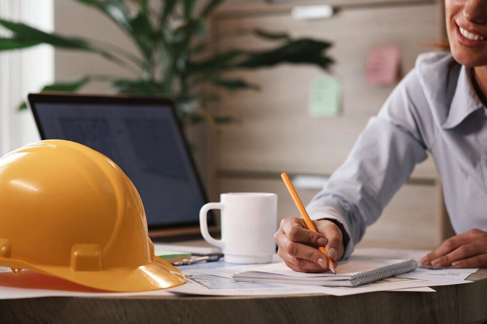 Worker writing beside a yellow hard hat, laptop, and coffee mug on a desk