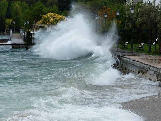 Large waves crashing over a stone seawall in a park during rough weather.