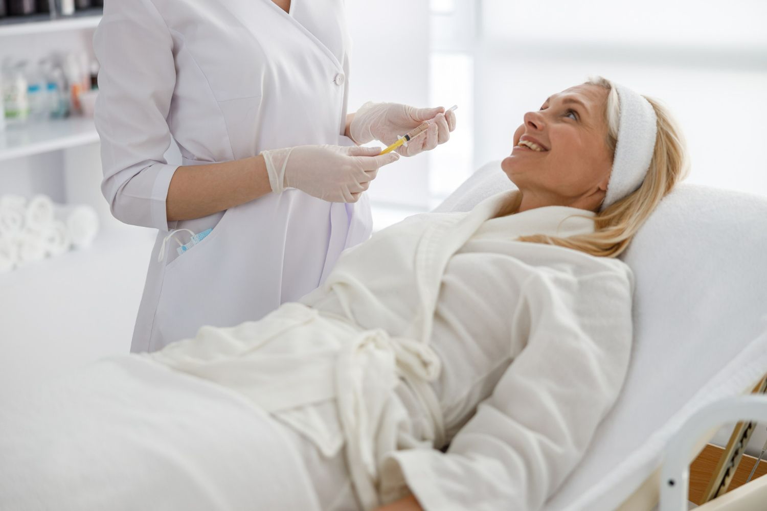 A medical professional in gloves holds a syringe near a smiling person in a spa robe reclining on a treatment table.