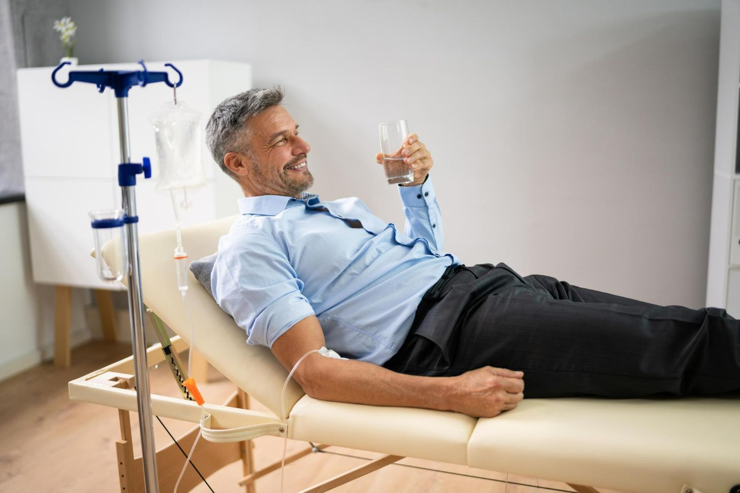 A person lying on a medical exam table while connected to an IV drip, smiling and holding a glass of water.