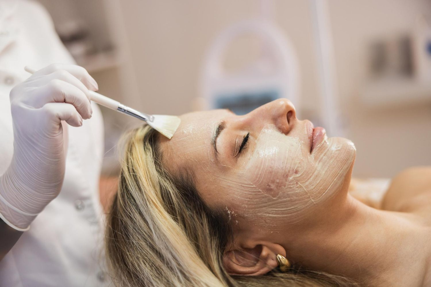 A person receives a facial treatment while lying down as a professional applies a mask with a brush.