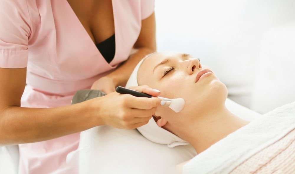 An aesthetician uses a brush to apply a white treatment mask to the face of a person lying down at a spa.