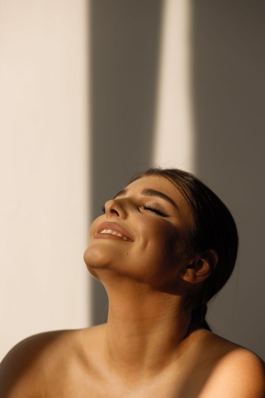 A woman is smiling with her eyes closed and the sun is shining on her face.