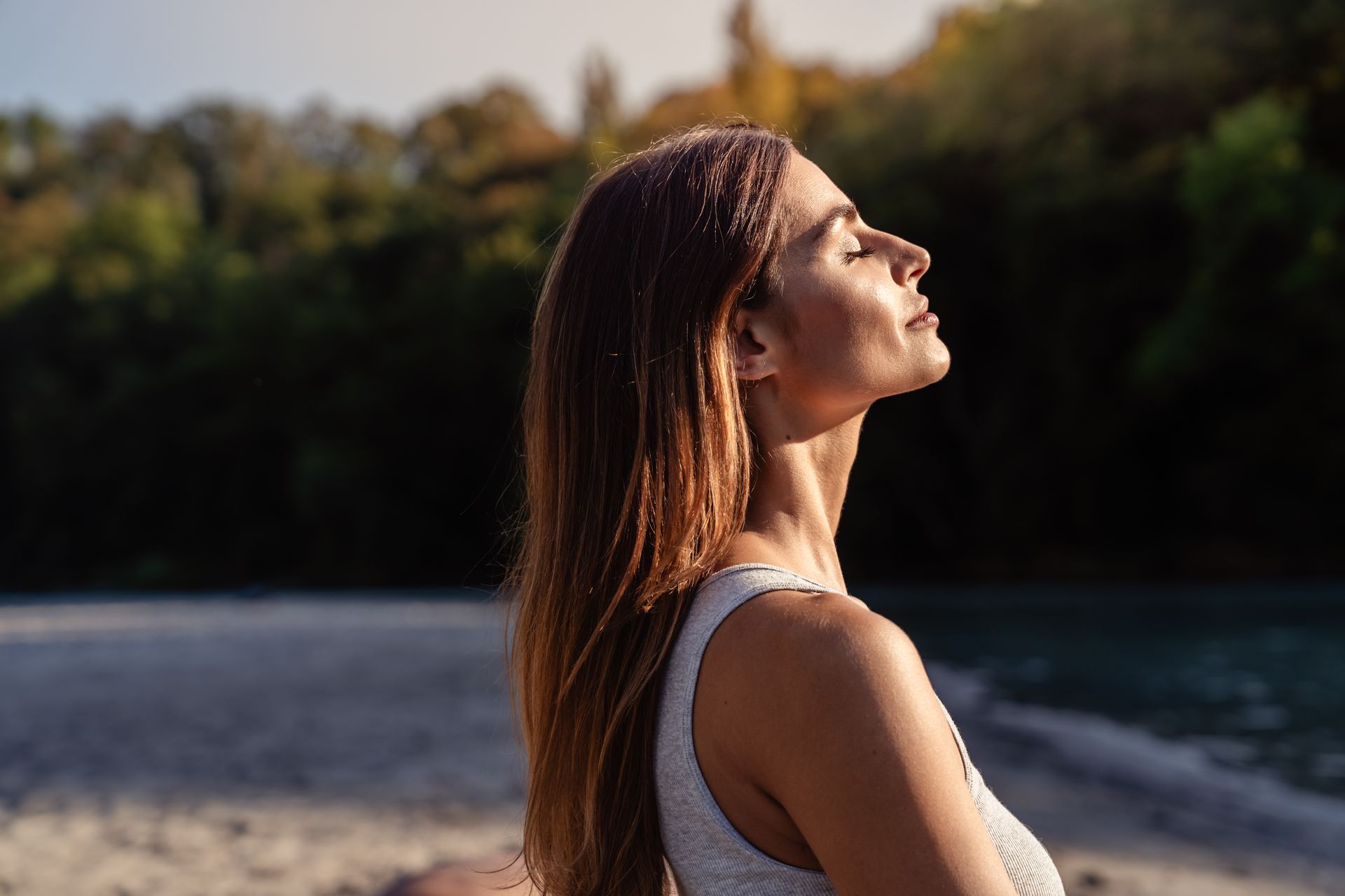 Woman in profile by a river, eyes closed, with sunlight on her face and trees blurred in the background