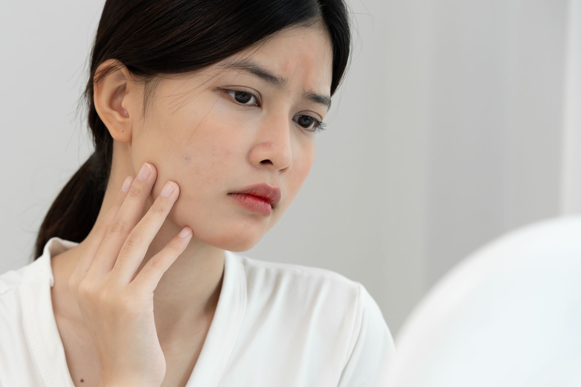 Woman looking at her face in the mirror, touching her cheek with a concerned expression. White background.